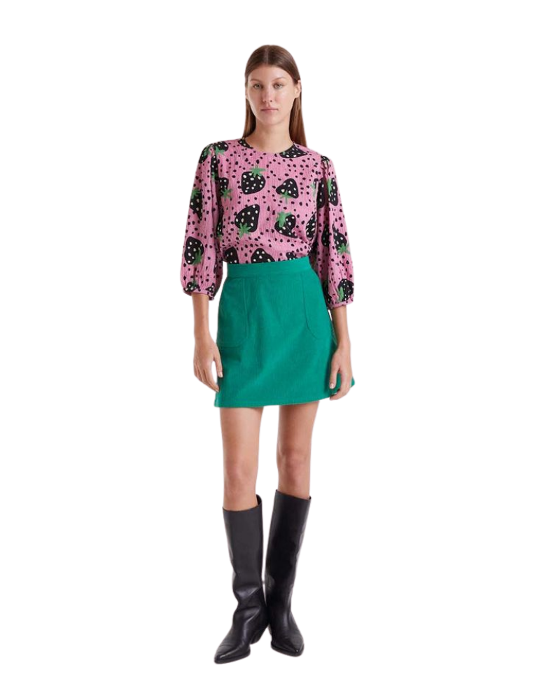 Woman wearing a pink blouse with strawberry pattern, green skirt, and black boots on a white background