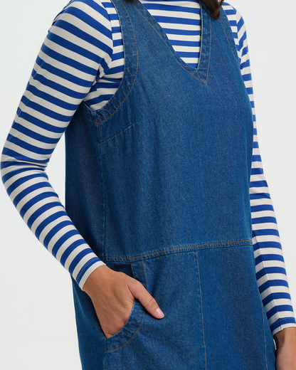 Person wearing a blue denim pinafore dress over a striped shirt on a white background