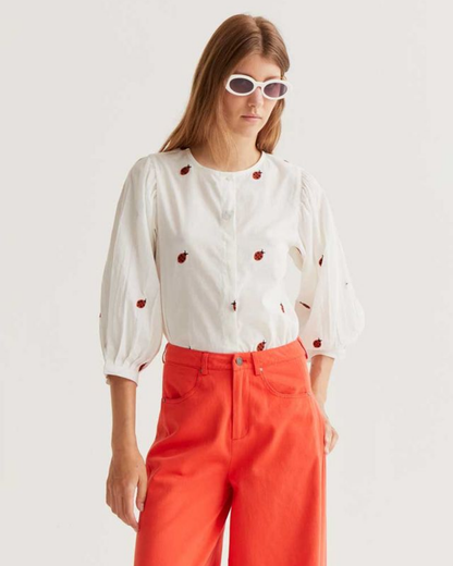 Woman wearing a white blouse with a ladybird pattern and red pants on a plain background